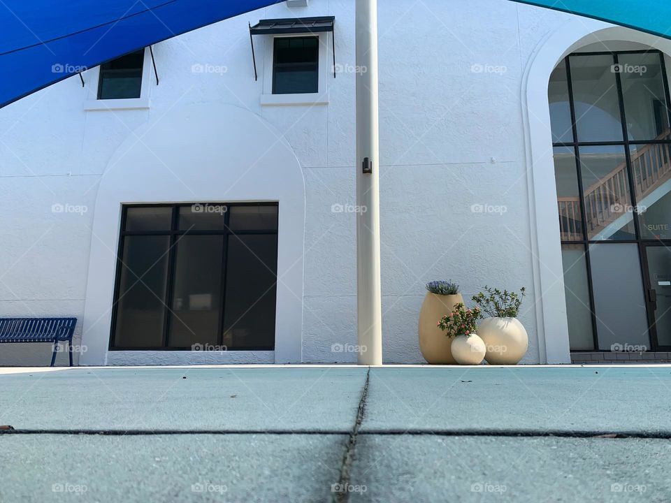 Architectural white and pure looking beautiful building with tanned windows and grilles windows with beige metal pole centered by three flower pots supporting blue and teal awnings.