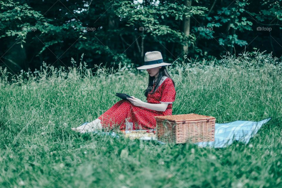 One young beautiful caucasian girl in a red dress and a straw hat sits on a bedspread with a wicker basket browsing social networks on a tablet while relaxing in a park on a summer day, close-up side view. Picnic time concept.