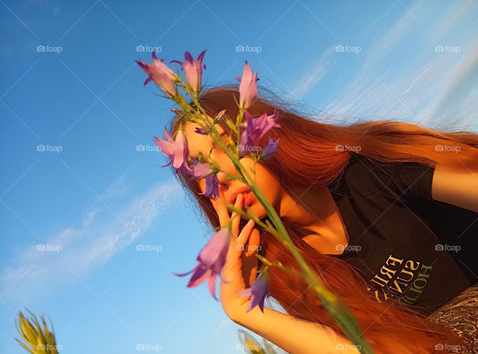 It is a photo of a young woman in the field. She has red (ginger) hair. She has brown eyes. Her face is partly coverd with Campanula patula. Her lips are full. She touches her lips with fingers