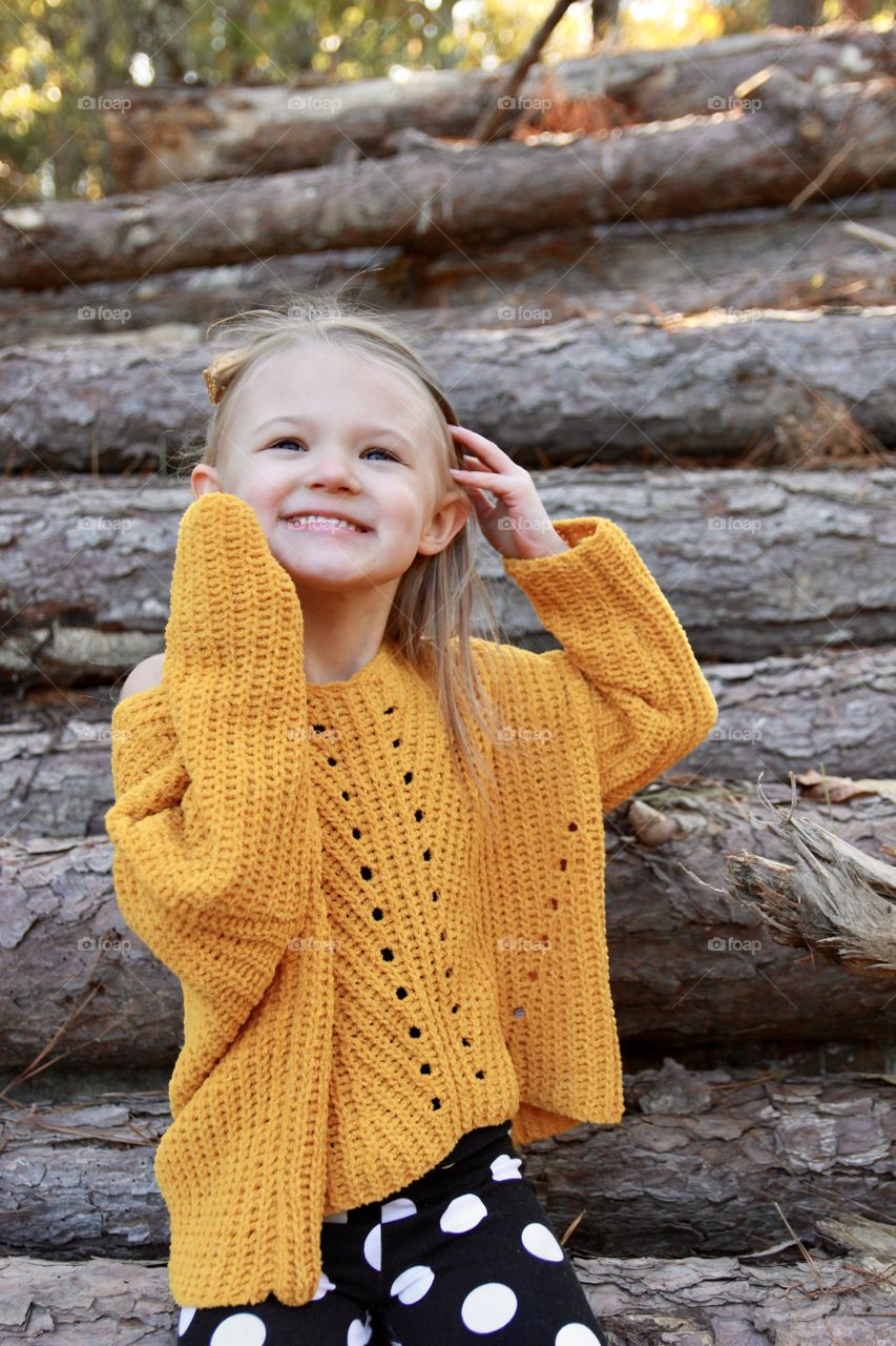 A little girl wearing fall fashions smiles and looks up. Behind her are stacked tree logs. 