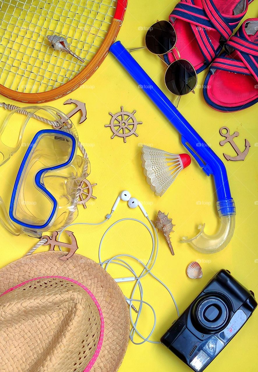 On a yellow background are items for traveling at sea: a straw hat, a camera, headphones, flip flops, sunglasses, a snorkel for diving, a mask for diving, a racket and a shuttlecock