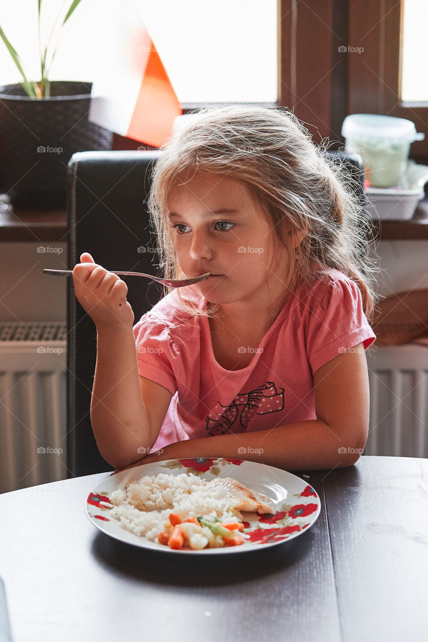 Little girl having a dinner at home. Meal on a plate on table, kid holding a fork. Real people, authentic situations