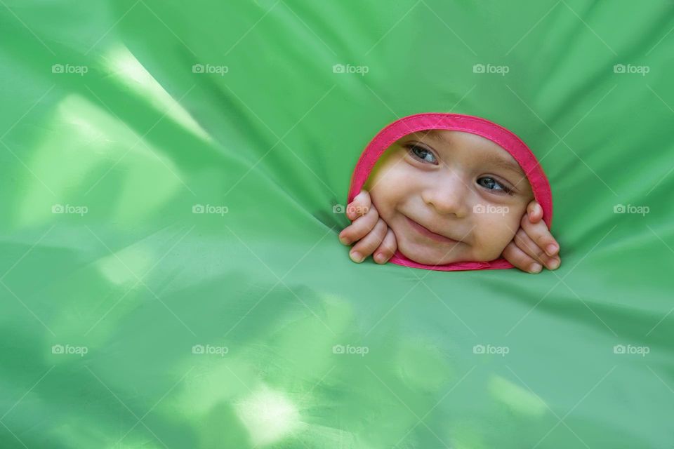 Portrait of happy smiling little girl looks out the hole of a green toy house with copy space. Background for advertising.