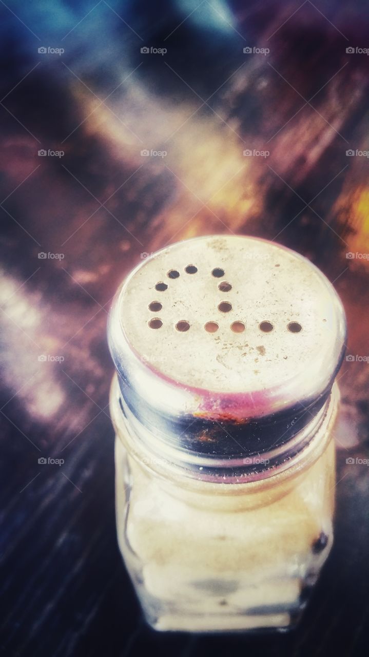 A pepper shaker on a wooden table with multi-colored reflections. Small round dots punched to the top of the shaker confirms it's content.