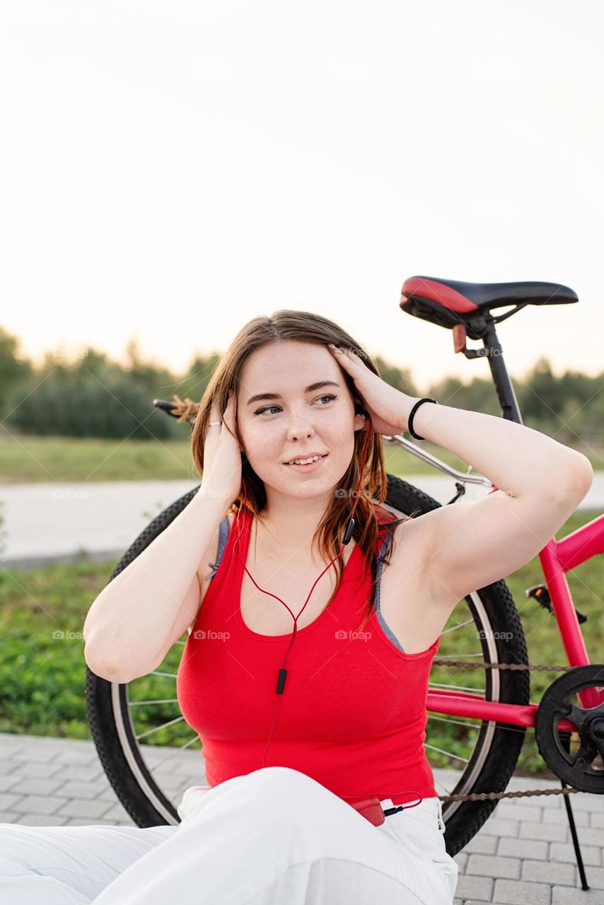 young smiling woman in red shirt and white pants listening to the music standing near her bicycle