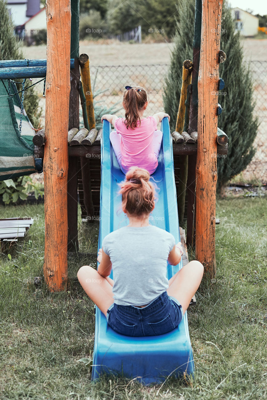 Teenage girl playing with her younger sister in a home playground in a backyard. Happy smiling sisters having fun on a slide together on summer day. Real people, authentic situations