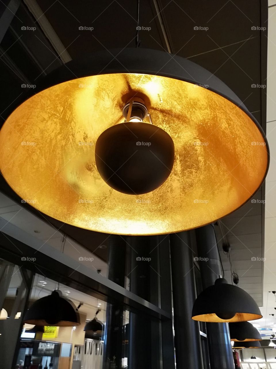 A large round lamp hanging from the ceiling in the restaurant. The surface of the light is matt black and inside it is yellow golden color with bright reflection, the black lid in the middle.