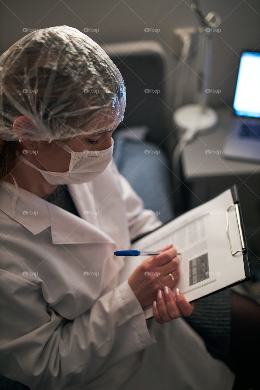 Doctor filling out a document. Hospital staff working at night duty. Woman wearing uniform, cap and face mask to prevent virus infection