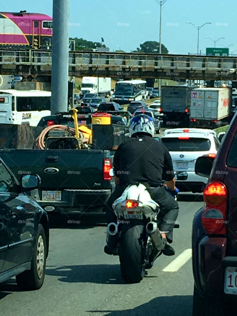 Subway trains, trucks, cars & a motorcycle all stuck in traffic.Thick traffic jam going home from work or going too work in the City.  There is also an overpass bridge for subway trains.