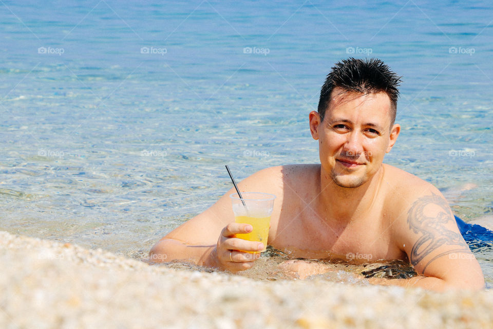 Man with a glass of cool fruit juice stay in water near the sea shore in the sunny summer day.