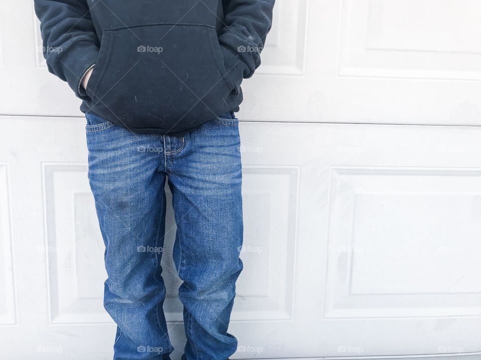 Boy standing next to wall wearing blue jeans and black hoodie with hands in his pockets 