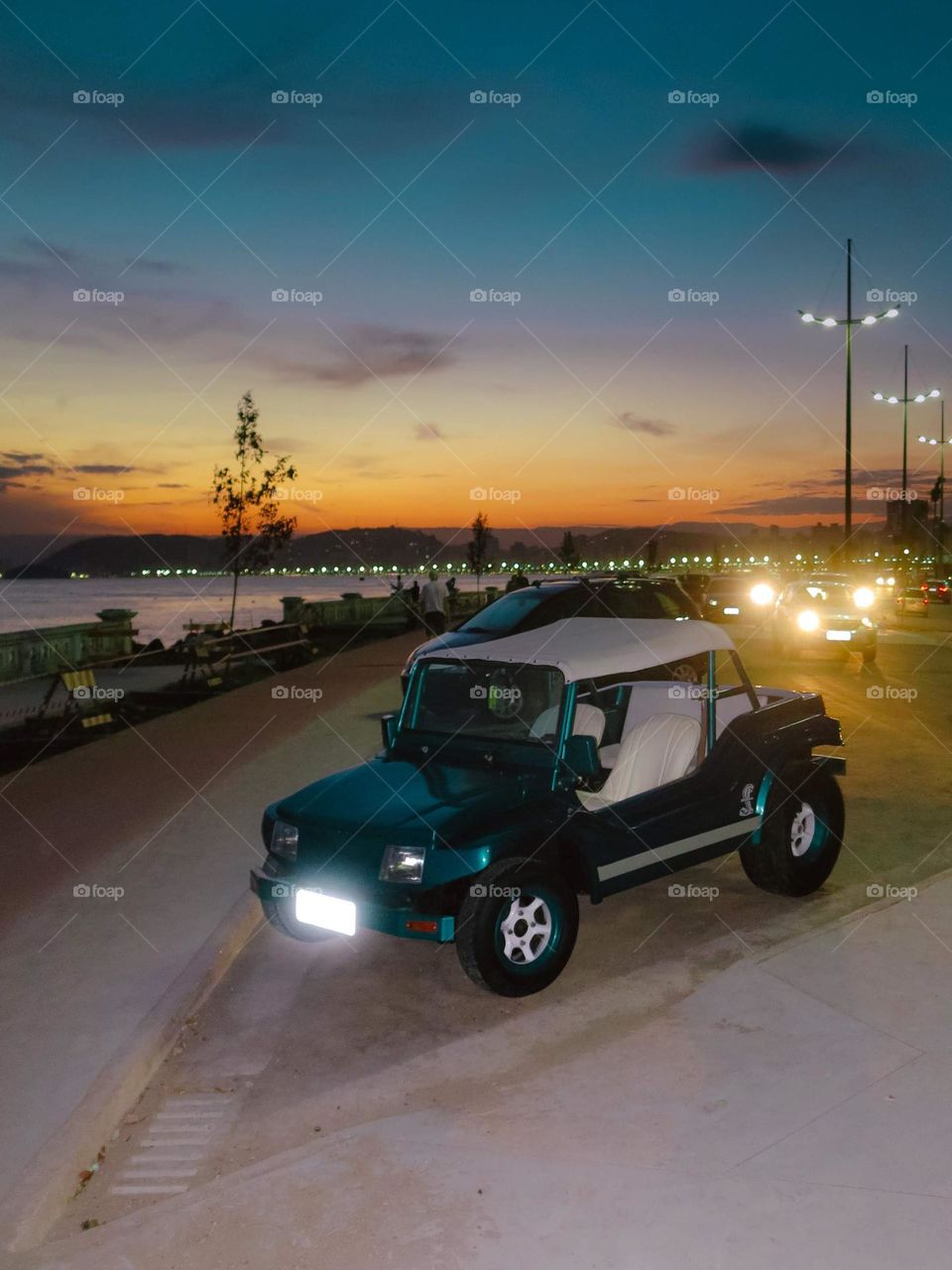 Green Buggy Car on the beach, at sunset. There's a busy street and a gigantic crate in the background.