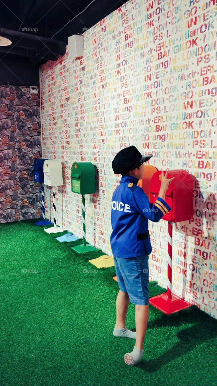 A boy in a blue shirt and hat walks towards a mailbox to send a letter. The colorful mural of letters and numbers on the wall adds a lively atmosphere, making the scene full of whimsy and imagination.