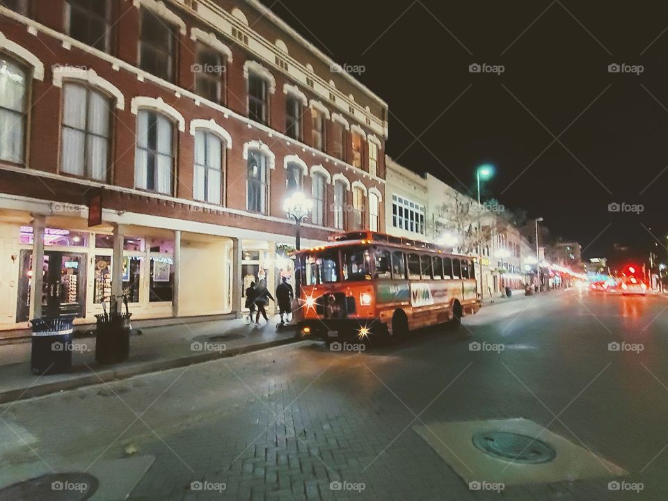 Downtown city trolly at night,  next to an old three story brick building with many windows.