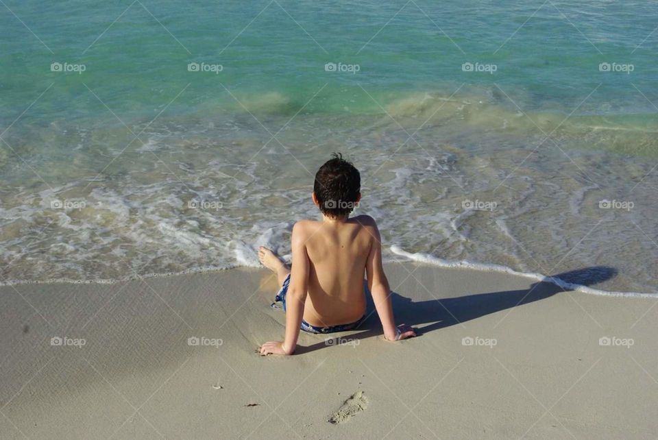 Young boy sitting on the beach, feet in the water.