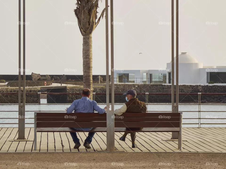 elderly man sitting on a bench talking in times of covid overlooking the sea