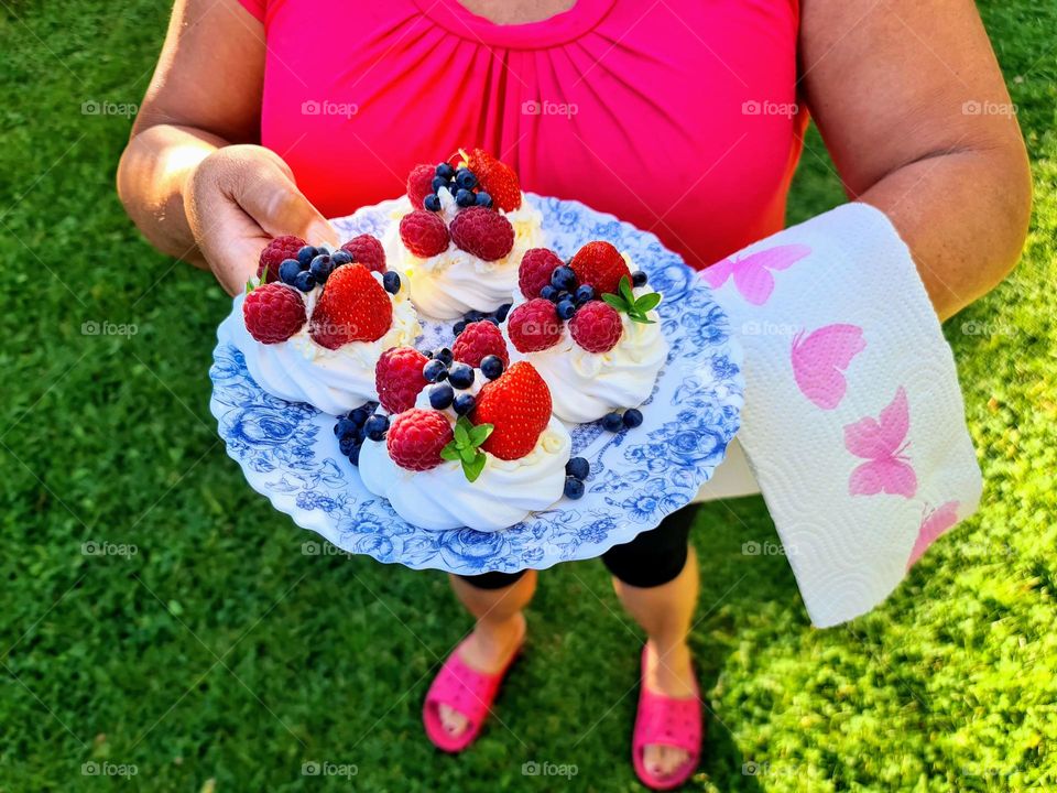 Delicious sweet meringue dessert with fresh berries strawberries, blueberries and raspberries on the blue and white plate held by a woman in the bright fuchsia top