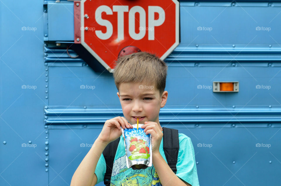 Young boy smiling and drinking from a drink pouch in front of a blue school bus