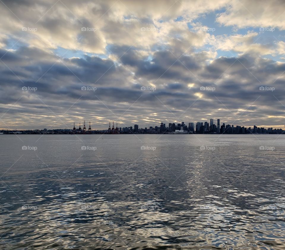 View from above, where sky meets ocean at Vancouver city skyline. Colors of grey and blue, in wavy ocean and cloudy sky, with silhouette of buildings in distance.