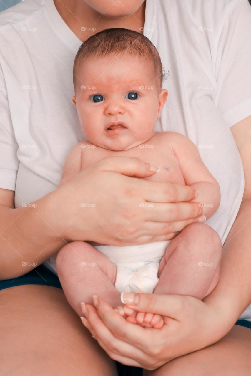 Portrait of a young mother tenderly holding her newborn baby while sitting on her bed during the day, from the side close up.