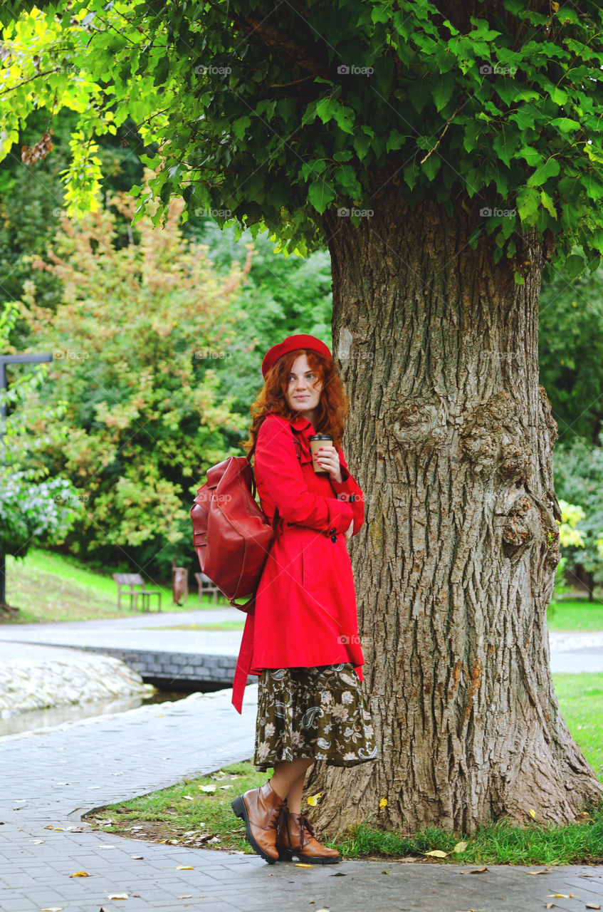 Young happy woman with red hair, freckles in a red beret and a coat walking in autumn park. Fall cold season.
