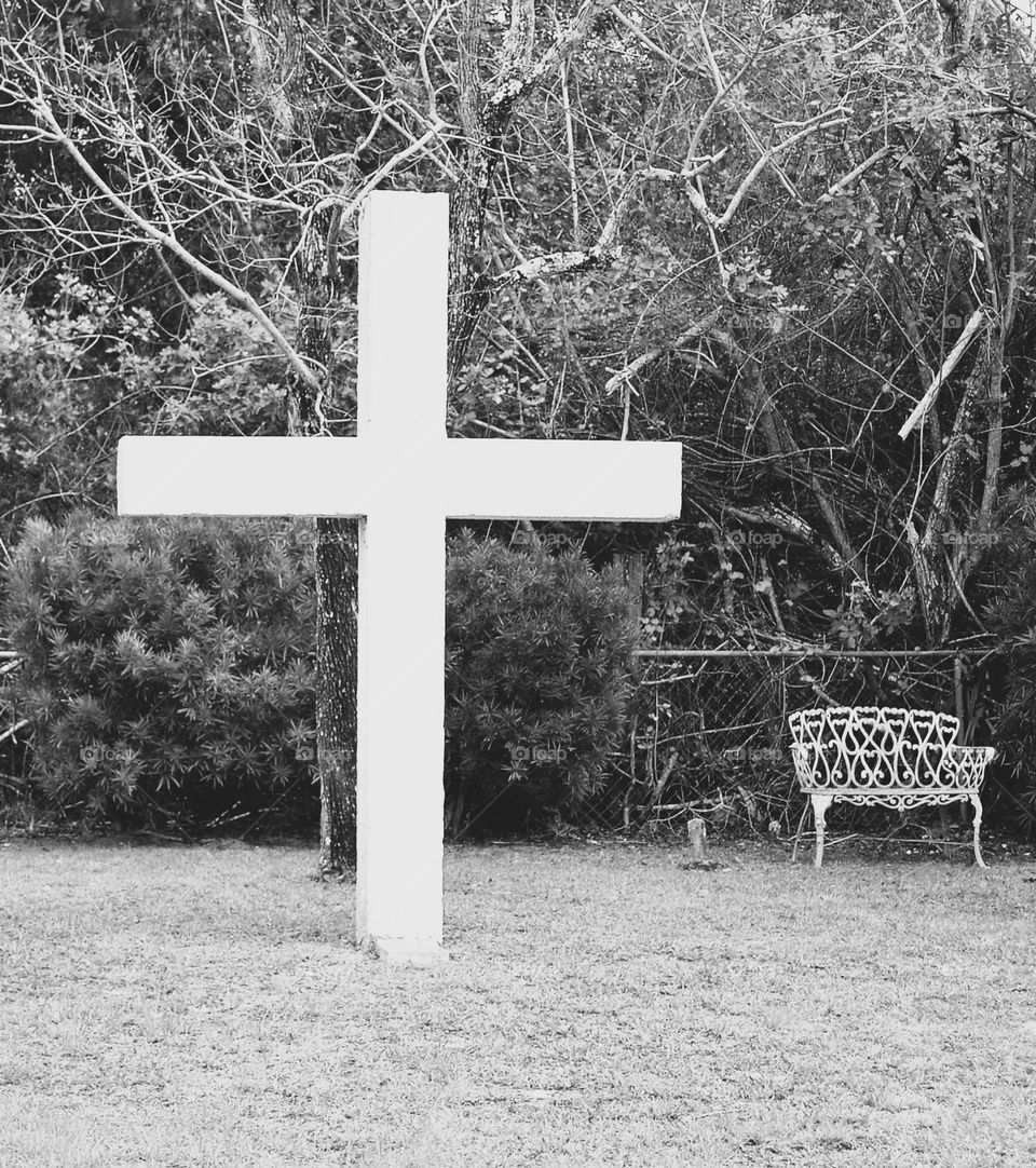 A black and white view of a large cross in front of bushes and trees and beside an ornate iron bench