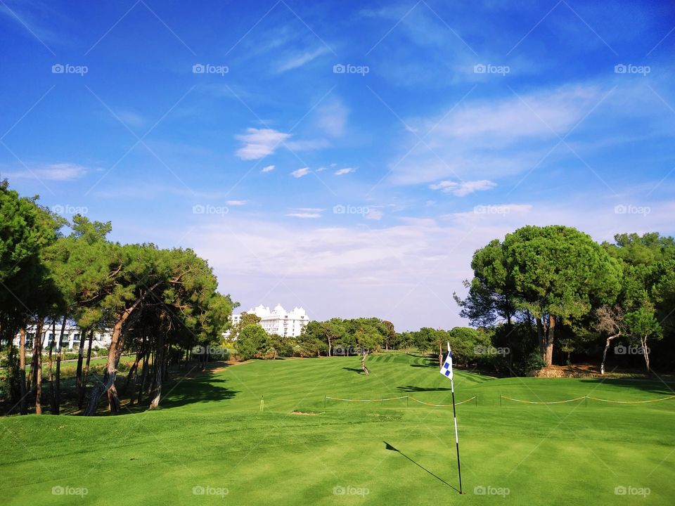 A beautiful golf hole view from behind the flagstick with daylight and shadows, trees, buildings and beautiful sky.