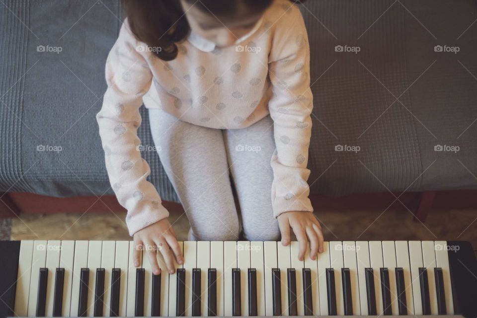 Portrait of a beautiful little caucasian brunette girl with two ponytails on her head plays the electric piano while sitting on the sofa in the living room, top view close-up. Music education concept, musical instruments,
children playing.