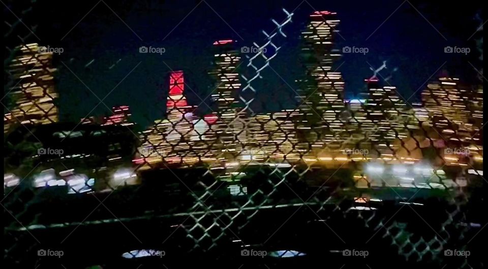 This is an interesting view from the pedestrian lane of the “Pulaski Bridge” at “Newtown Creek”. Through the illuminated grating of the fence LIC and further back “Manhattan”, even the “Empire State Building” can be seen. 2023. Hypnotic Productions