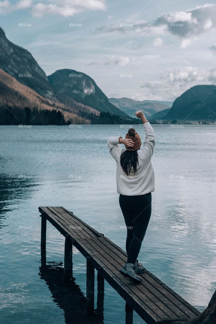 Rear view of young woman standing on small woode pier on shore of a lake in mountains on a moody winter day