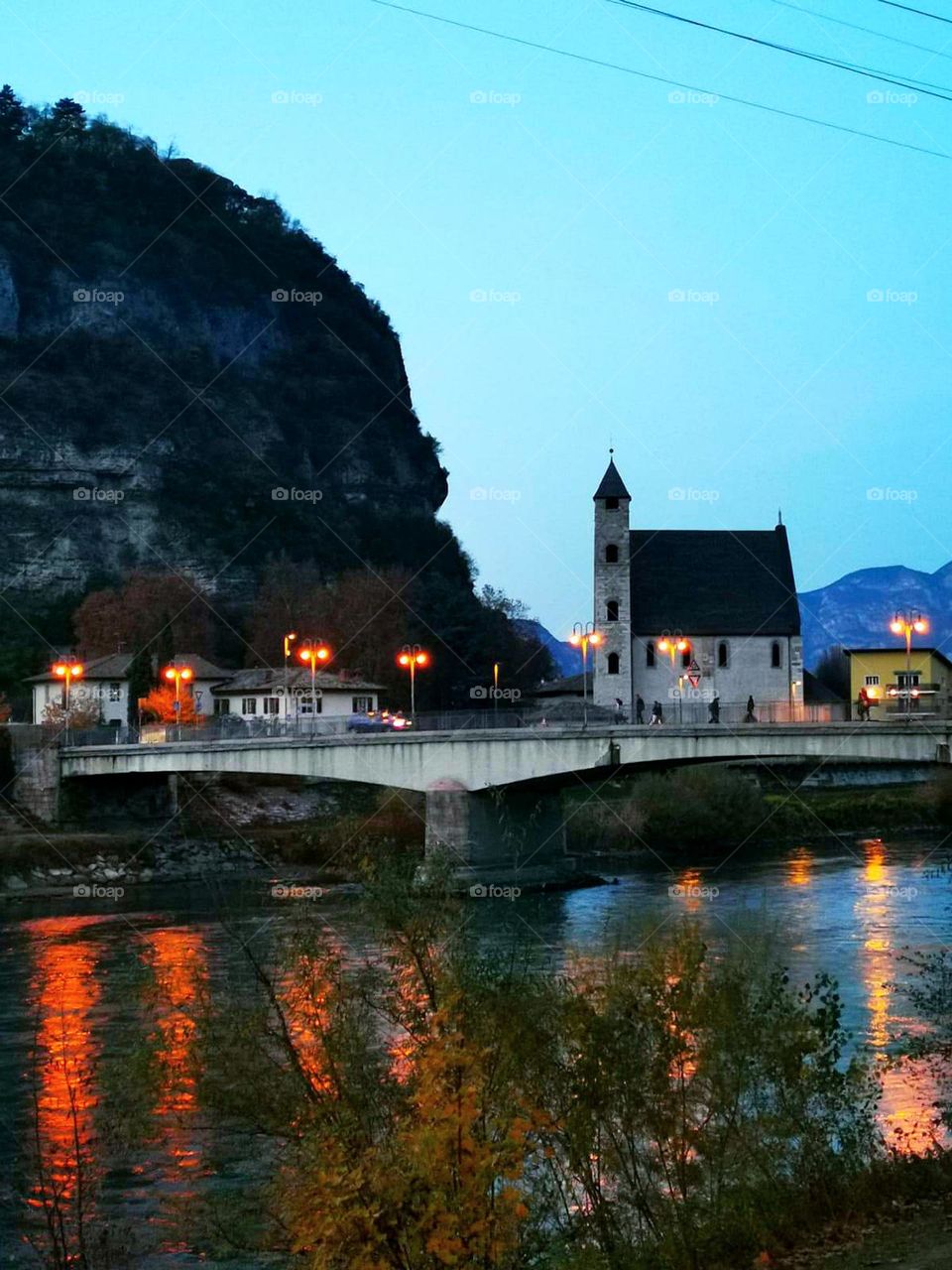 Evening. View of the city of Trento with a mountain in the background. The city between the mountains and the river. The church and the glowing street lights are reflected in the river. Italy