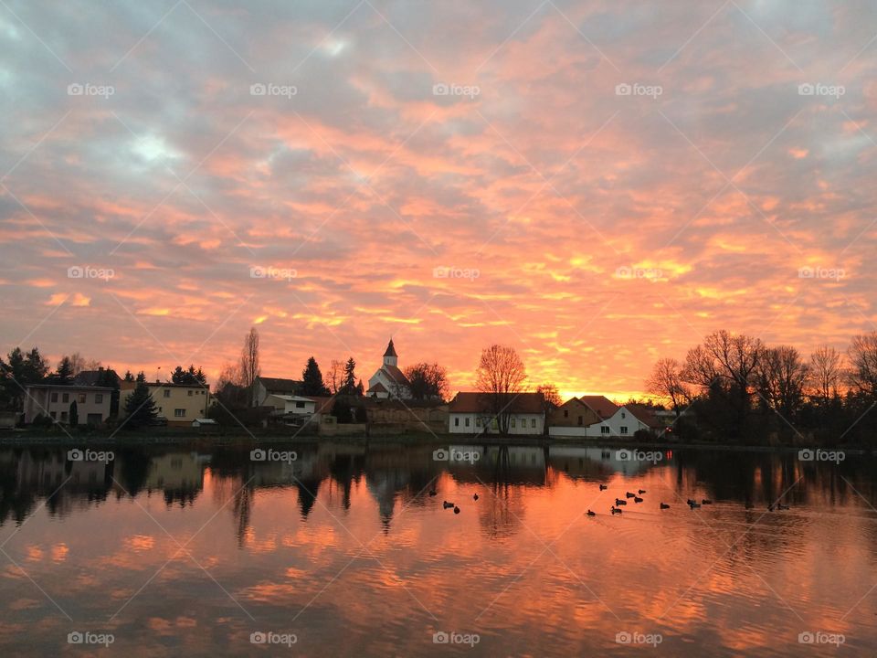 A beautiful sunset reflected in the pond of Hovorcovice village. View of the village with the church. The setting sun and the sky full of red colored clouds.