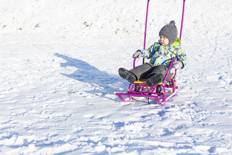 A child with a serious expression on his face in winter clothes jackets, pants, hat and boots in winter on white snow on the street and in the park in nature sledding and playing winter fun.