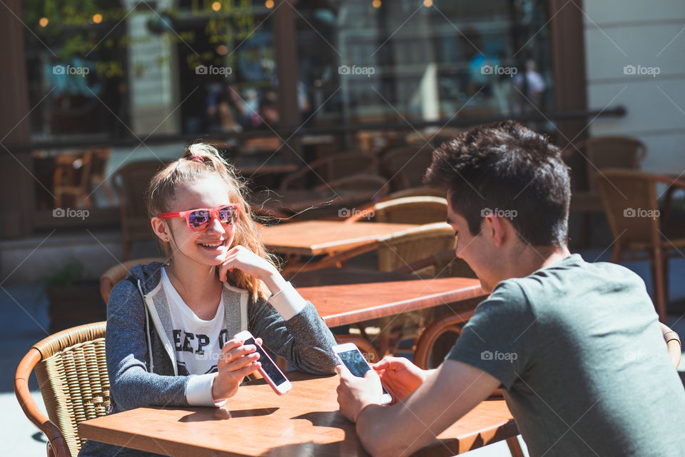 Young woman and man sitting in pavement cafe a the table talking and using mobile phones