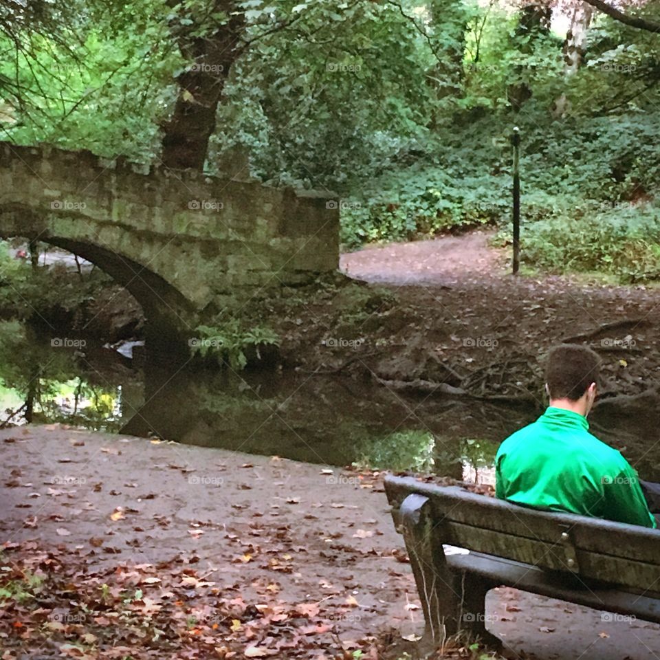 A man sat reading by a river over a bridge. 