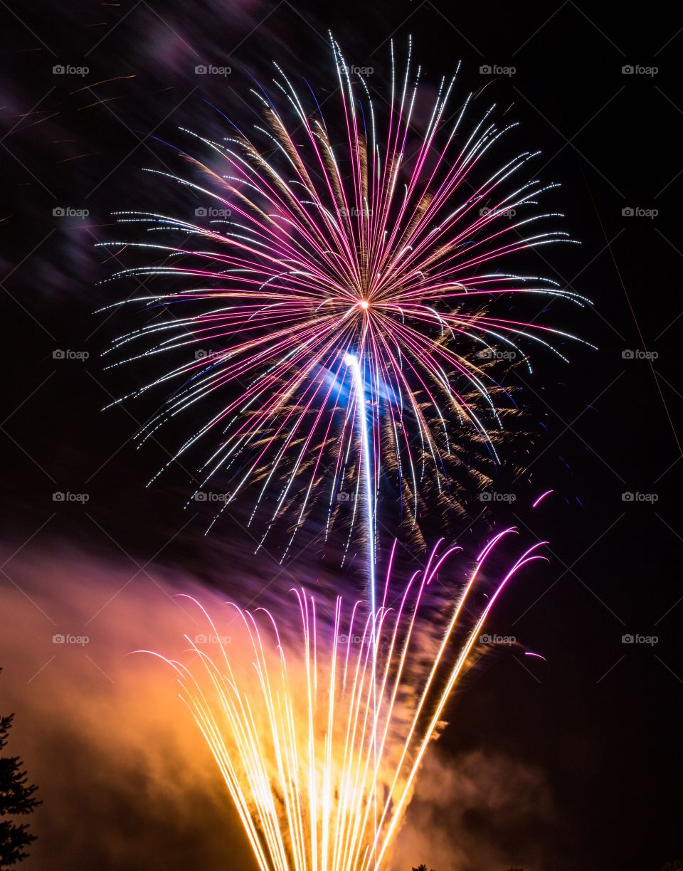 Vertical photo of yellow, orange and purple fireworks shooting up from the ground while a multicolor firework explodes in the night sky