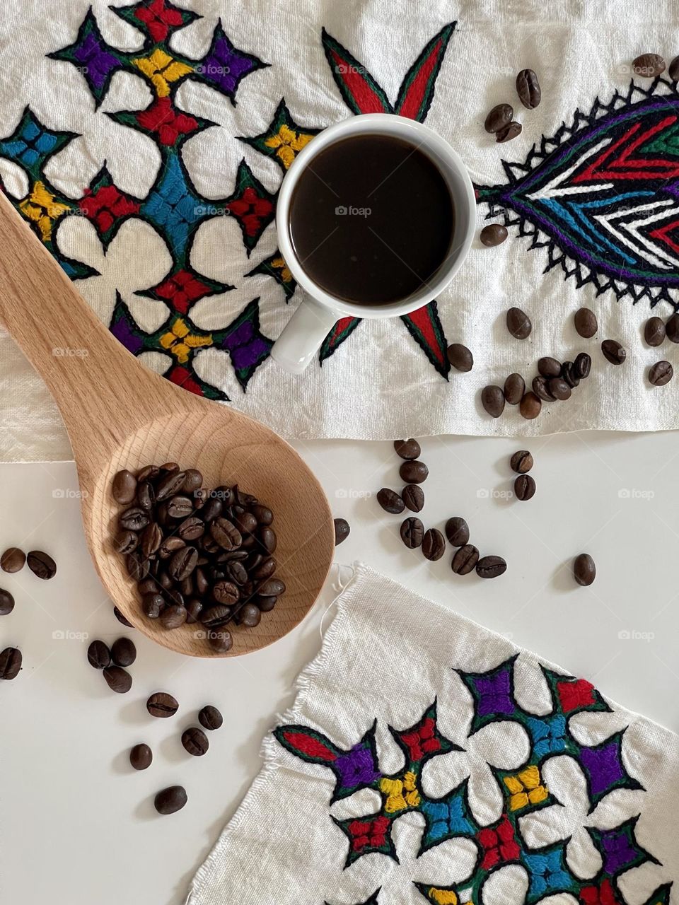 coffee theme flat lay: white coffee cup containing liquid coffee and coffee beans on white surface and inside a natural wooden spoon, on hand-embroidered Eritrean drape with typical ethnic embroideries