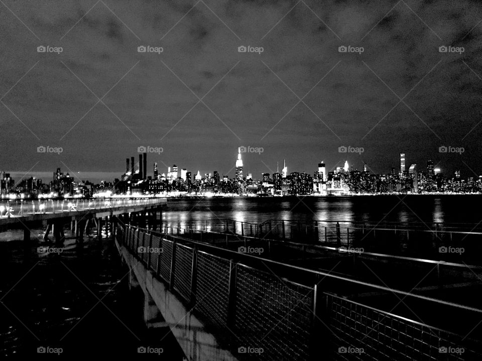 Black and white of the Manhattan skyline seen at night from Brooklyn. Clouds are seen in the sky and building reflections on the East River. Empire State Building is seen. 