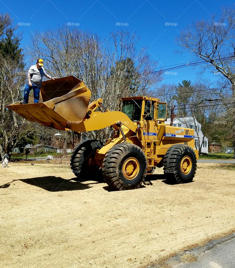 Front End Loader machinery on lawn doing work. Man standing in bucket, sunny day to work outside with heavy equipment.🚜