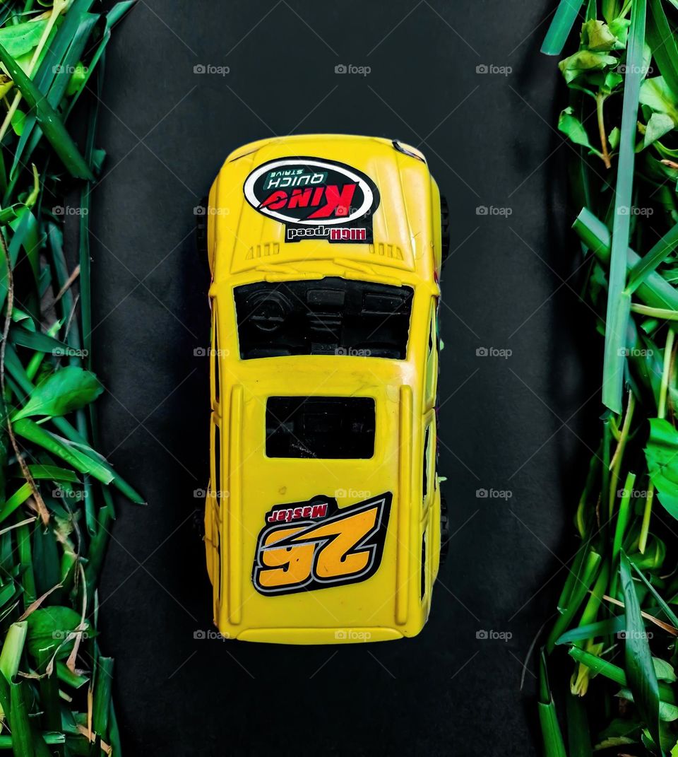 yellow toy car, seen from above, between a road and trees, made of grass