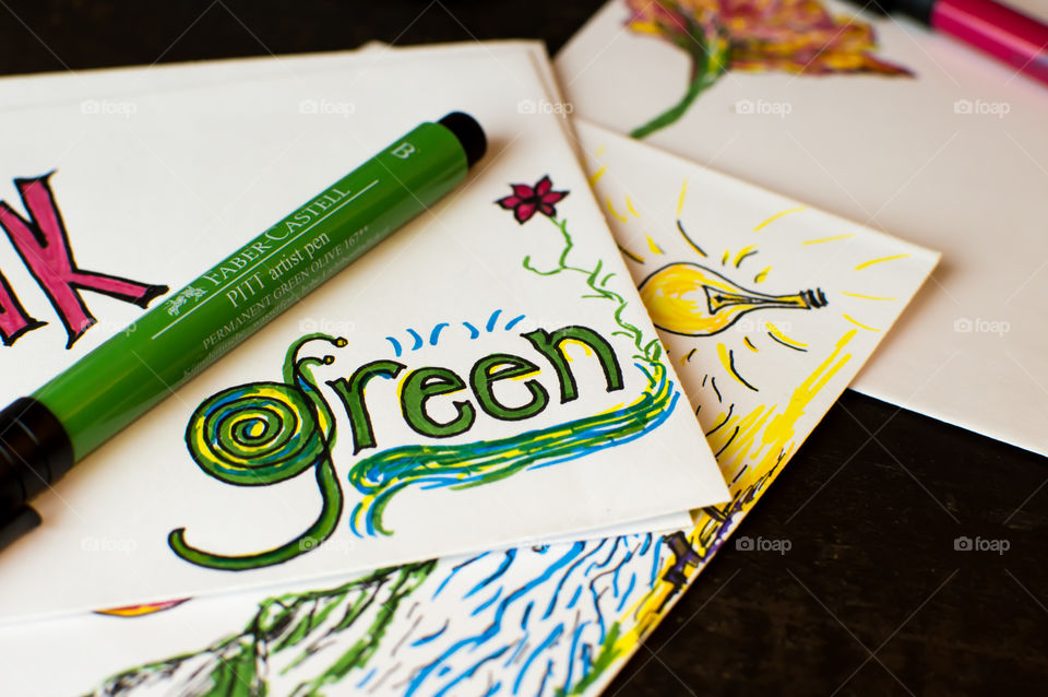Faber-Castell artist pen in olive green on desk with word “green” written in nature design on card stock next to light bulb a creative images conceptual ideas, earth day, green planet environmental words and images photography background