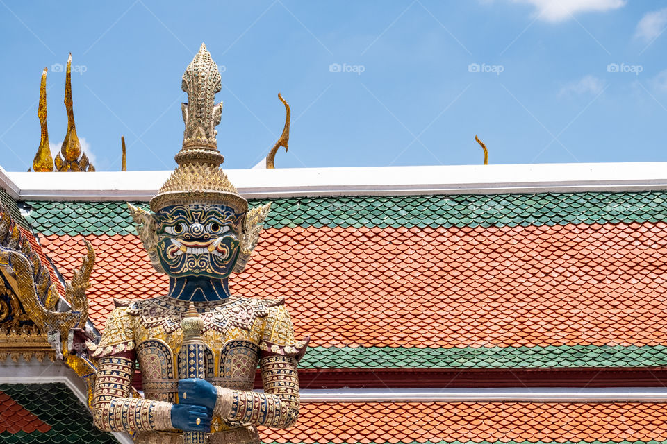 Giant statue in front of the brightly colored roof of Wat Phra Kaew