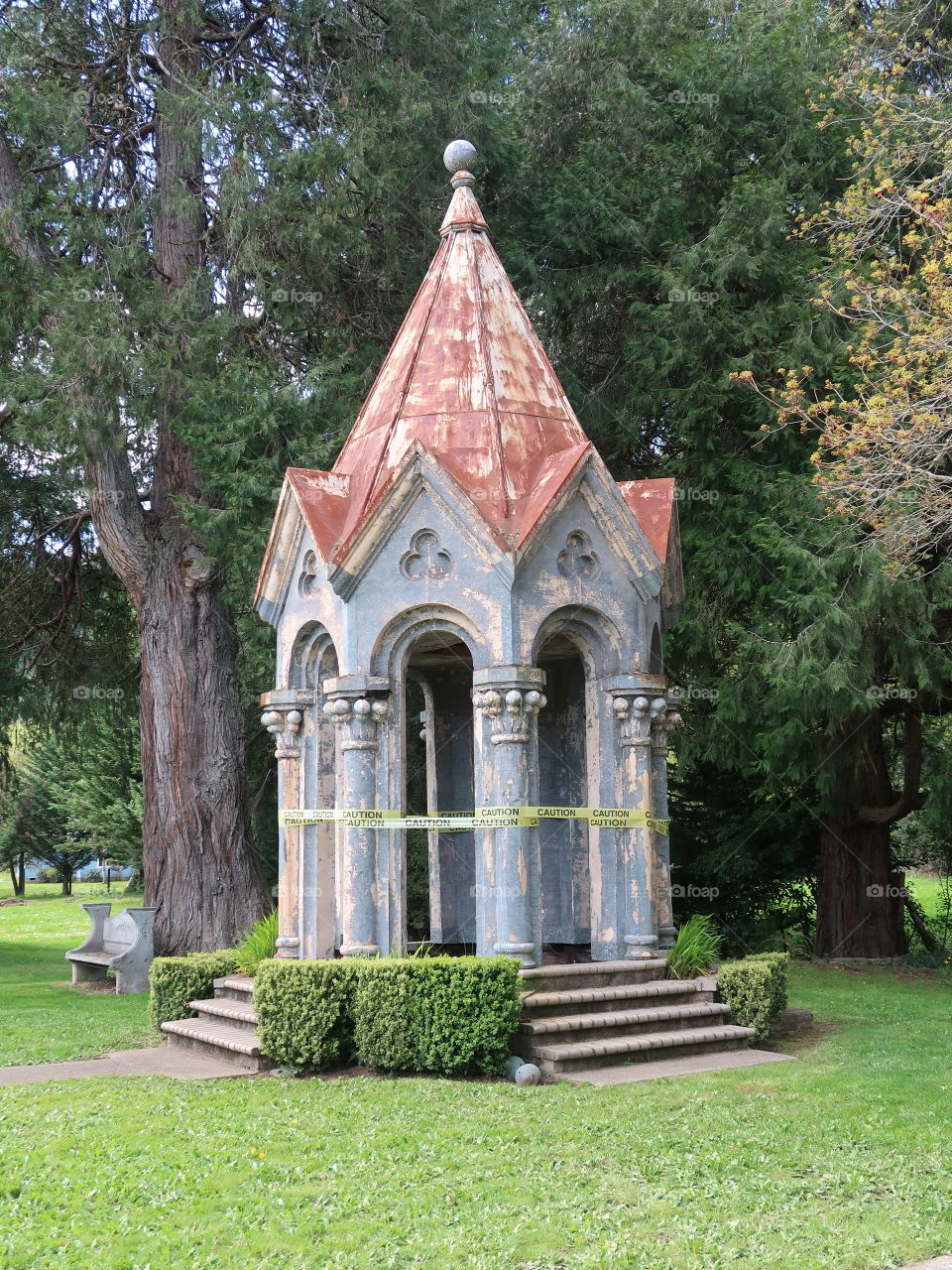 A large decorative stone gazebo in a nice park that has developed a lean and is in need of repair. 