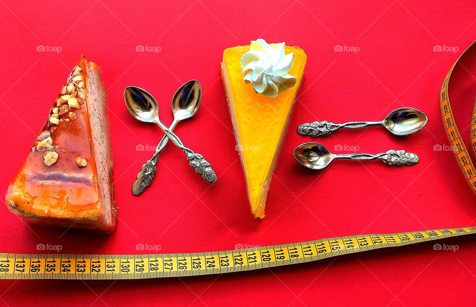 Triangles.On a red background lies a triangle of almond cheesecake and a triangle of lemon cheesecake.Between the cheese cakes are two crossed spoons. Behind the lemon cheesecake are two spoons in the shape of an inverse proportional sign.