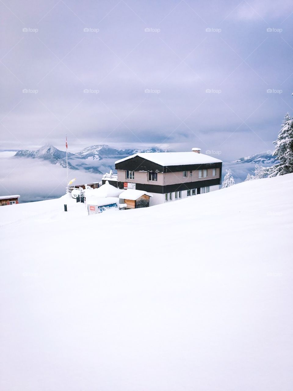 White snow covered the top of the mountain, a house in the middle, around the trees also covered with snow. Seeing a few mountains in the distance, clouds covered the sky, with no sunlight.  winter is beautiful but too cold