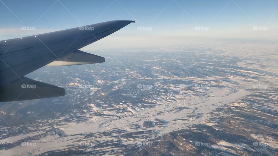 View from above, looking out of airplane window while flying over snow covered Canadian rockies during sunny day.