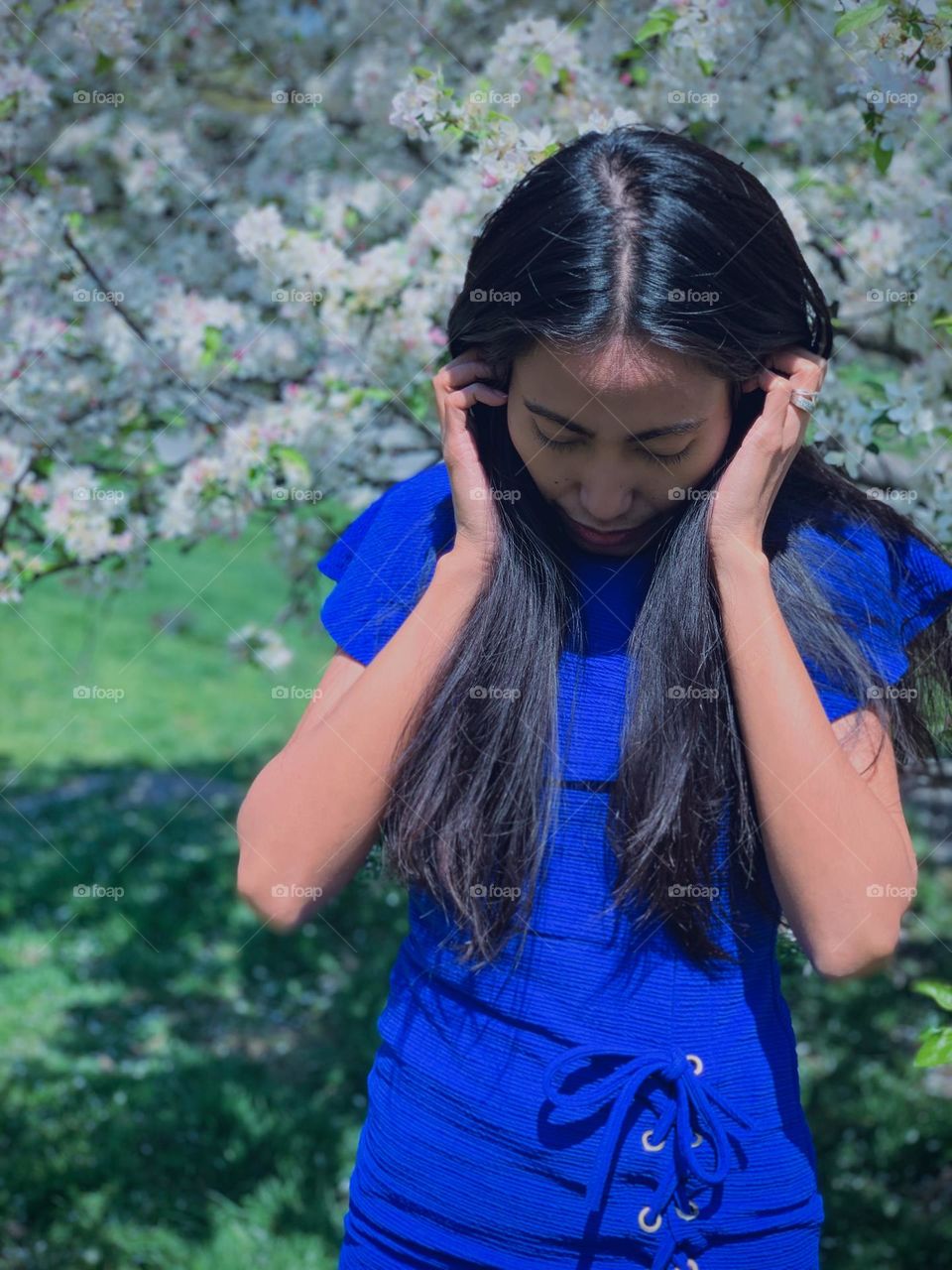 Portrait of an adult woman wearing blue casual dress, holding her hair and looking down against the backdrop Cherry blossom trees. 