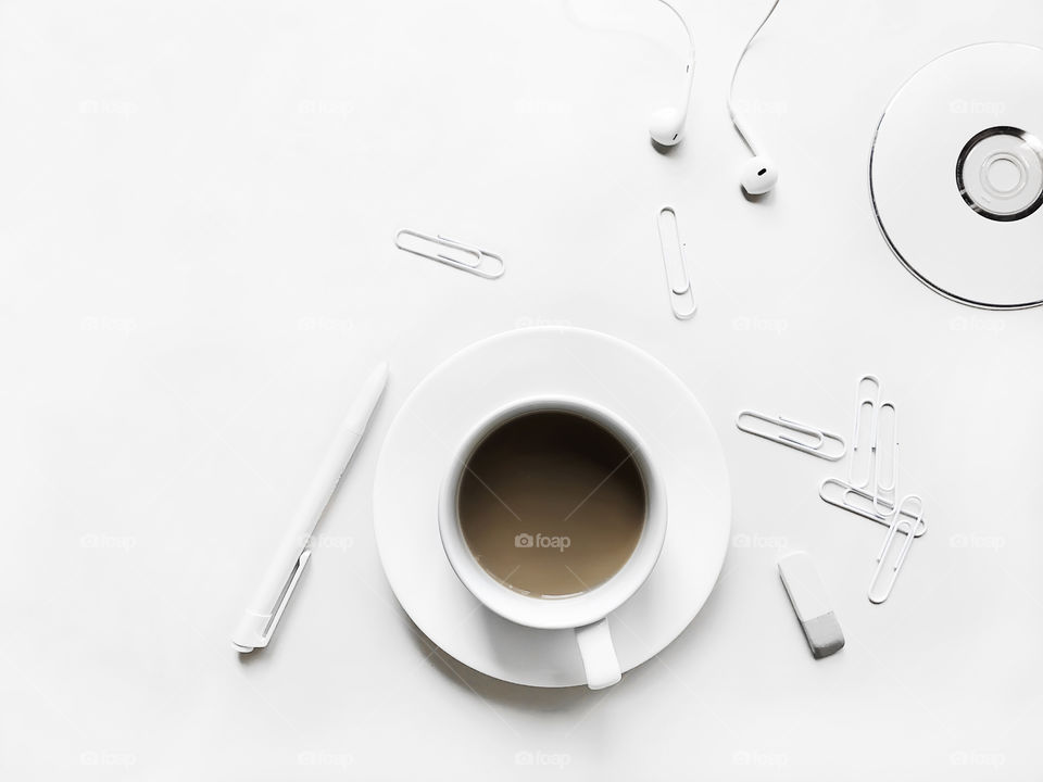 Coffee with milk in white cup, white office supplies and white headphones on white table 