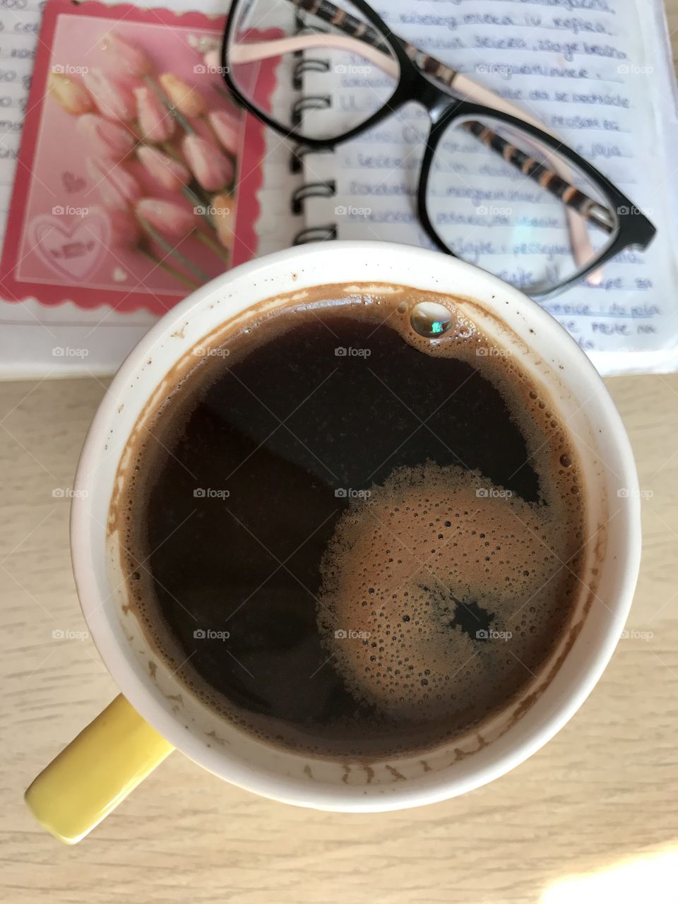 Yellow cup of coffee on the light wooden table with black coffee in it and with notepad, glasses and little rose pink picture of tulips flowers.