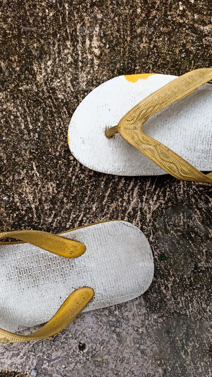 a pair of white flip-flops with yellow straps placed on a black and white surface. This photo depicts the striking color contrast between the flip-flops and the floor, as well as signs of wear on the flip-flops.